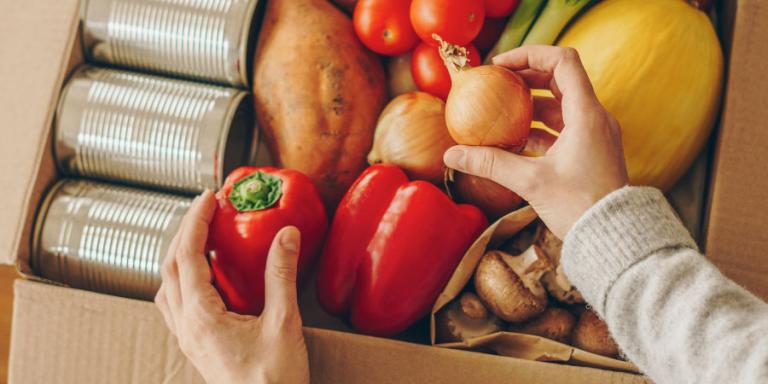 a pair of hands reaches into a box of produce and canned goods.
