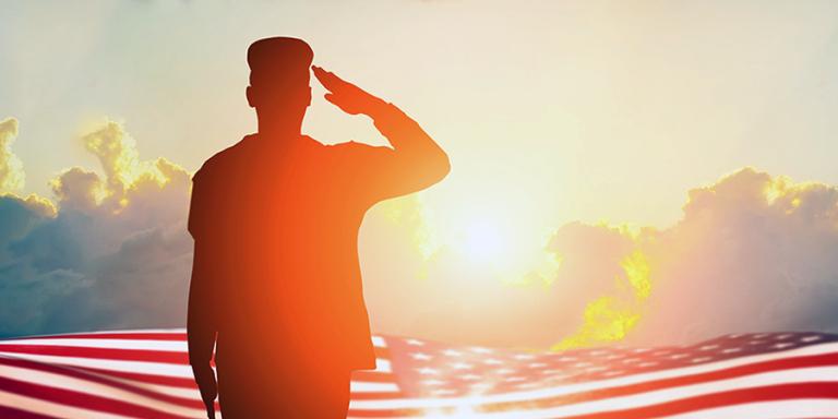 A veteran in shadow solutes the flag in front of a sunset 