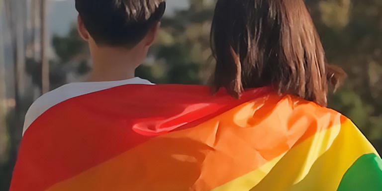 Two persons wrapped in a colorful rainbow flag celebrating Pride Month.